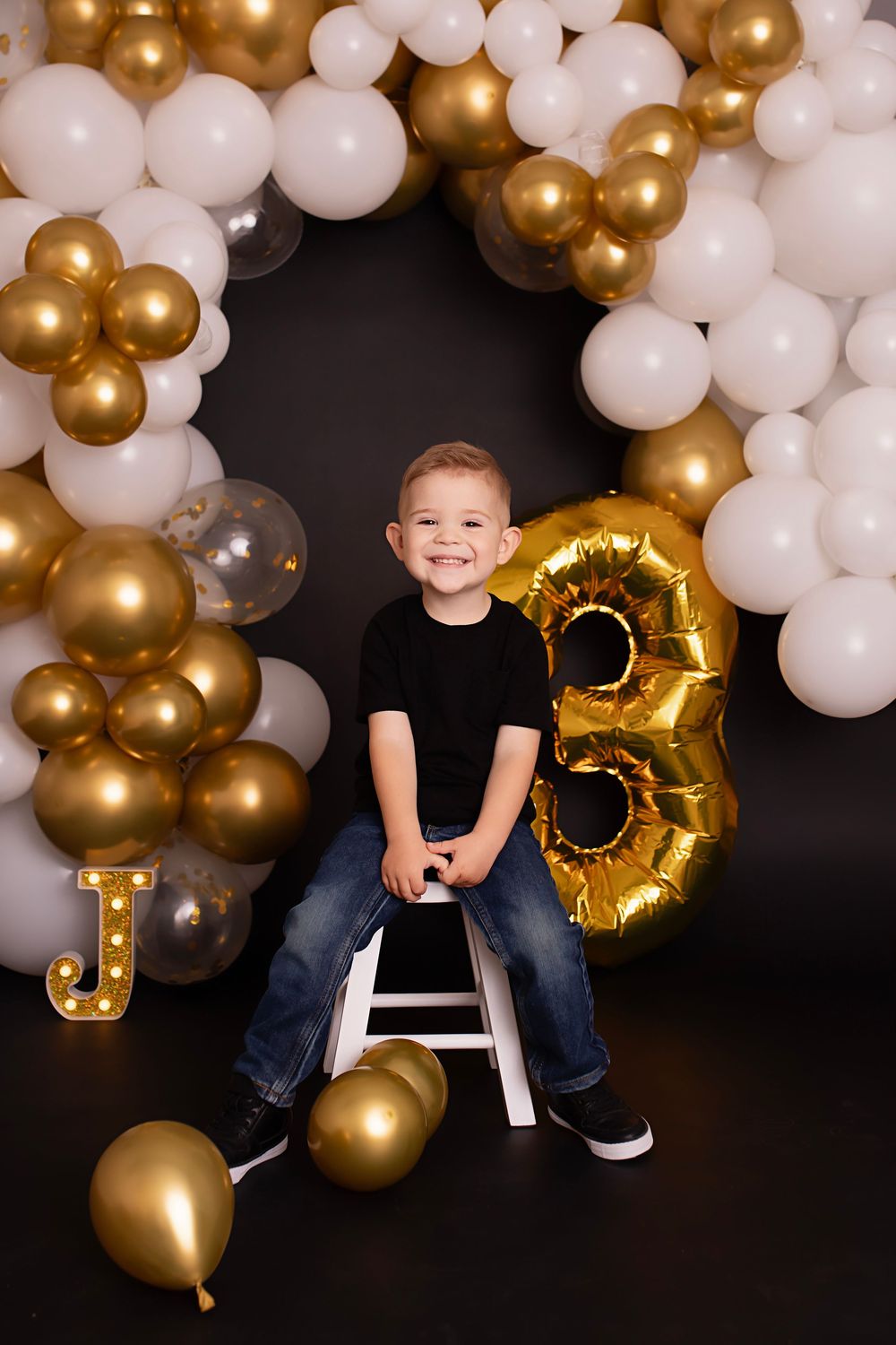 Black and gold balloon arch creates festive backdrop for birthday photo shoot with child on white stool.