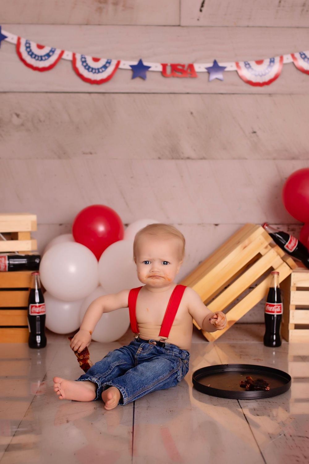 Baby in jeans lying on wooden floor with red and white balloons and patriotic bunting decorations in background.