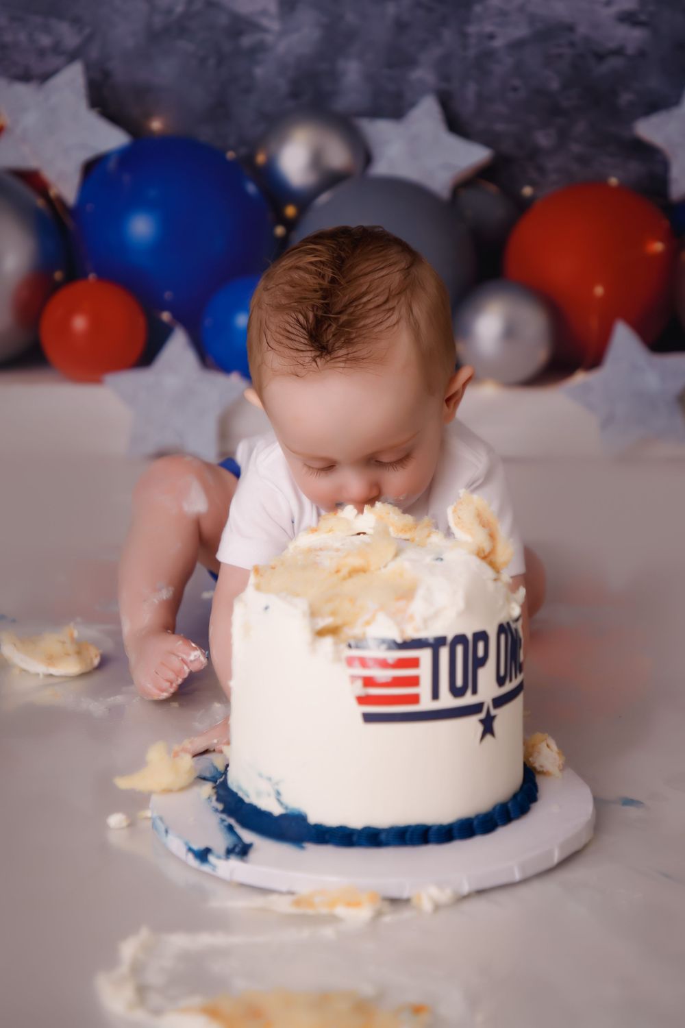 Baby smashes into a patriotic Top Gun themed cake with red, white and blue balloons and stars in the background.