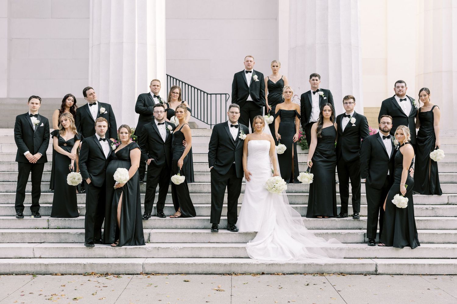 A wedding party in formal black attire and a white wedding gown poses on grand white stone steps.