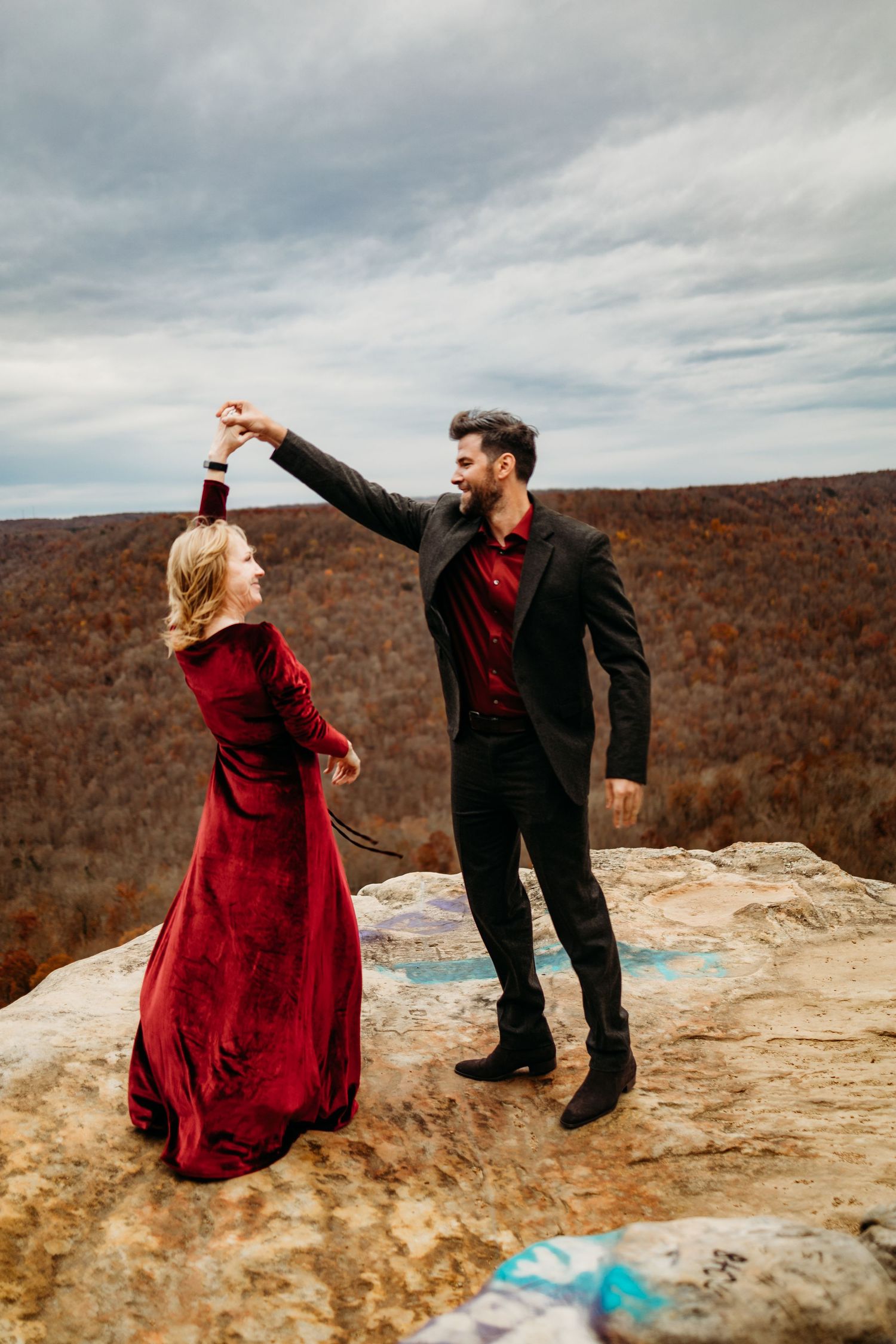 Man in black suit twirls woman in red velvet gown on mountain cliff overlook.
