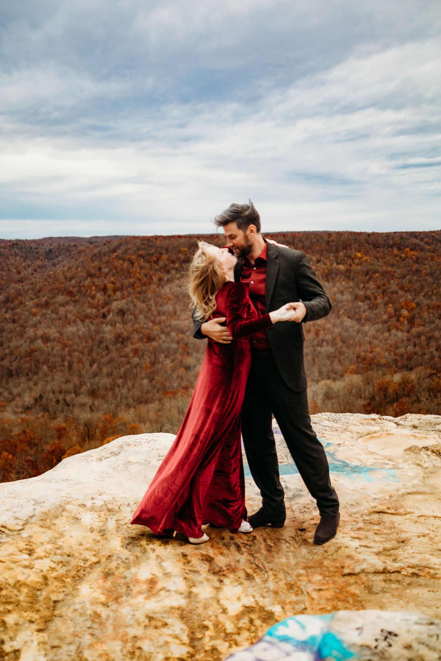 Couple in red and black formal attire embrace on rocky mountain summit in autumn.