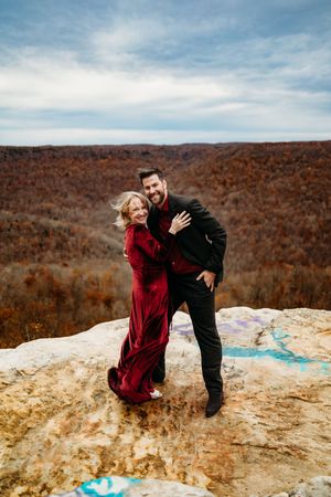 Couple embracing on rocky overlook in autumn, woman in red velvet gown and man in black suit with fall foliage backdrop.