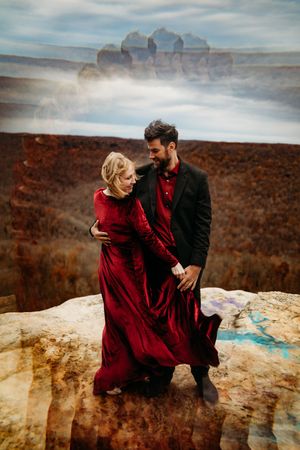 Couple holding hands on cliff edge in red and black formal wear with dramatic canyon landscape in double exposure effect.