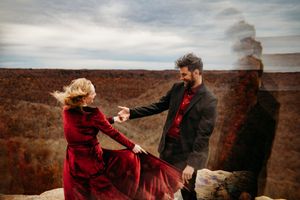 Couple dancing on rocky cliff with autumn forest backdrop, woman in flowing red velvet dress holding mans hand.