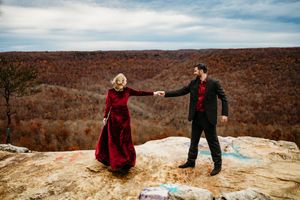 Couple holding hands on graffiti-covered rock ledge with vast autumn forest valley stretching into the distance.