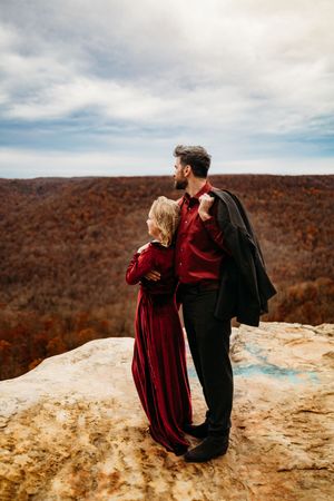 Couple standing together on mountain overlook, man in red shirt holding jacket over shoulder with woman in red gown.