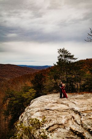 Couple embracing on dramatic rocky cliff edge surrounded by pine trees and colorful autumn mountain landscape.