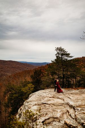 Couple kissing on rocky mountain overlook with pine trees and expansive autumn valley landscape under cloudy sky.
