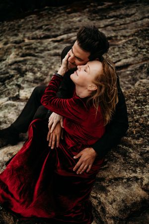 Romantic couple in red velvet dress and black suit sitting on rocks sharing an intimate almost-kiss moment.
