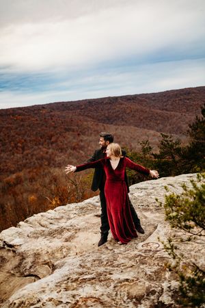 Woman in red velvet dress spreading arms wide on rocky summit with man in black suit and autumn hills behind them.