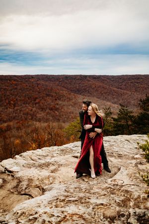 Couple in red velvet dress and black suit embracing on rocky mountain overlook with autumn forest valley backdrop.