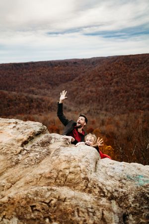 Joyful couple peeking over rocky cliff edge waving hands with colorful autumn mountain landscape behind them.