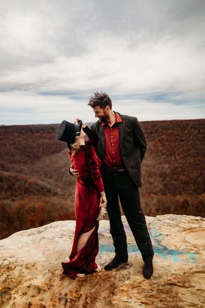 Couple in vintage red velvet dress and black suit sharing a playful moment on rocky mountain overlook in fall.