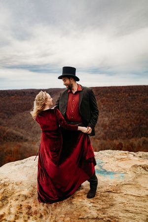 Couple in Victorian-style red velvet dress and black top hat dancing on rocky mountain cliff in autumn scenery.