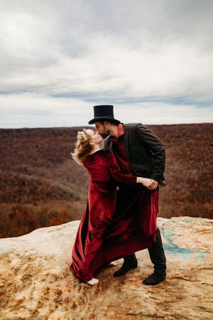 Couple in red velvet dress and black top hat sharing a romantic kiss on rocky mountain overlook in fall foliage.