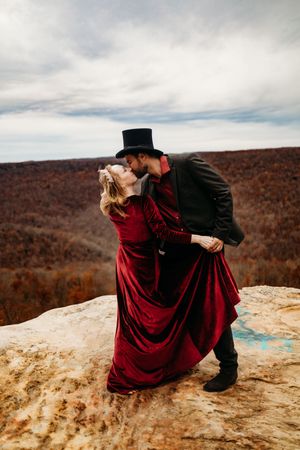 Couple in red velvet dress and black top hat kissing passionately on a rocky cliff with autumn mountain backdrop.