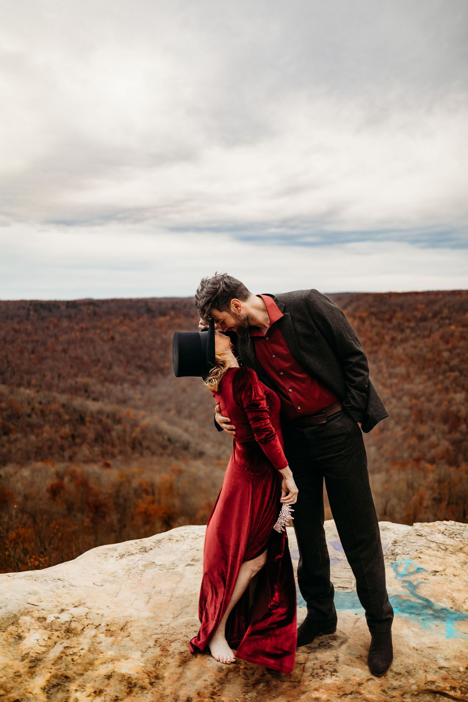 Couple in red velvet dress and black top hat sharing a deep kiss on rocky mountain overlook in fall landscape.