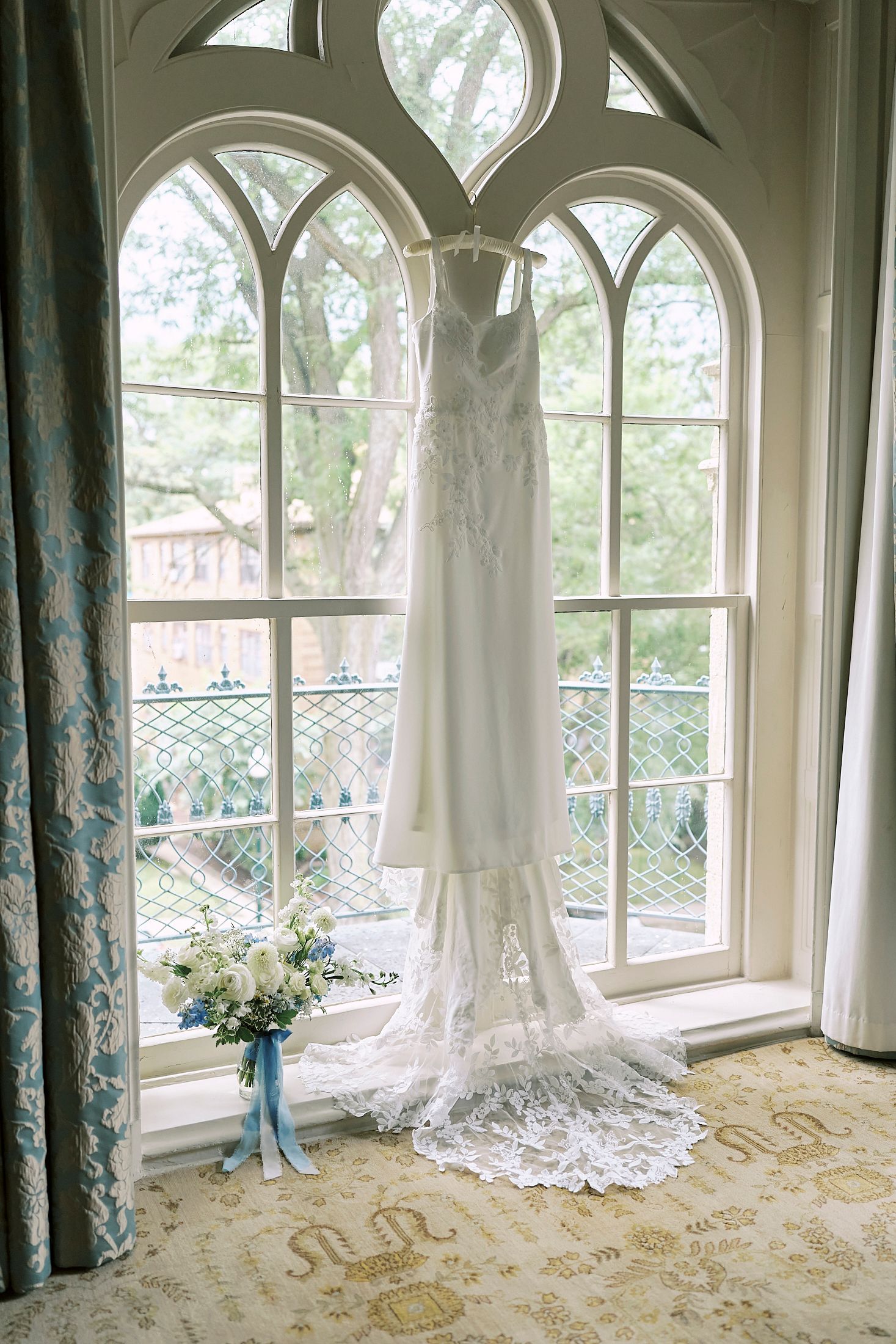 White lace wedding dress hanging in front of elegant arched windows with natural light streaming through.