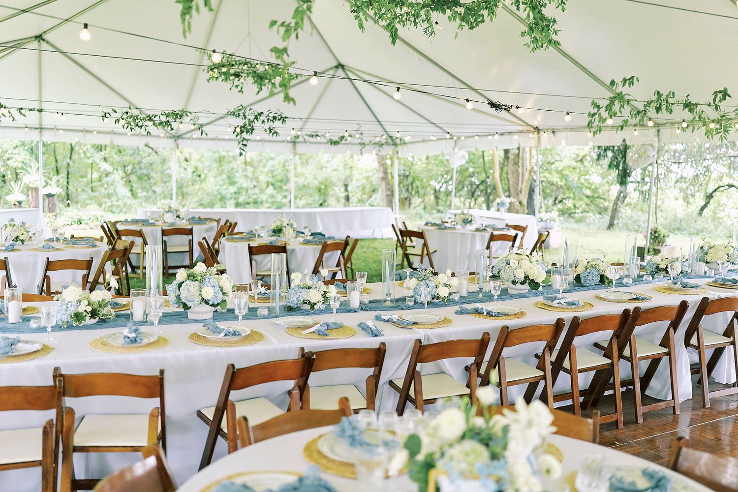 Long rows of wooden chairs and tables set up under a white tent with hanging greenery for an outdoor wedding.