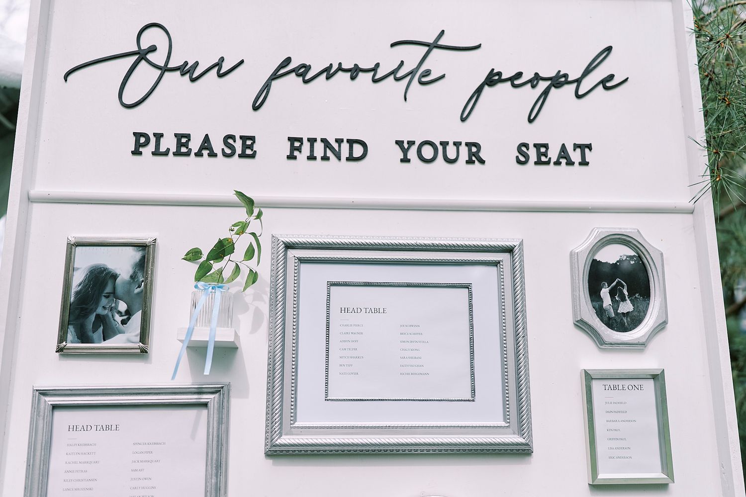 Wedding gallery wall setup with framed pictures, seating chart signs, and greenery decorations against a white wall.