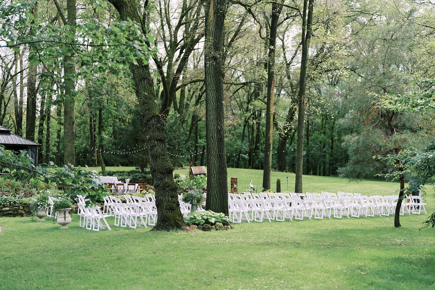 White chairs line a grassy outdoor ceremony space surrounded by tall trees in a garden setting.