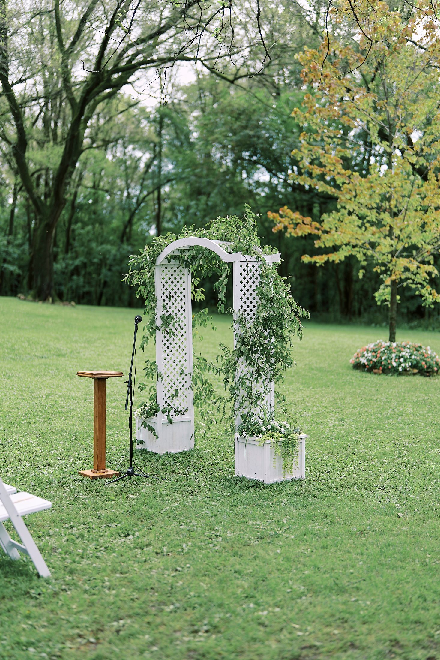 White lattice wedding archway with delicate floral arrangements stands elegantly on a lush green lawn surrounded by trees.