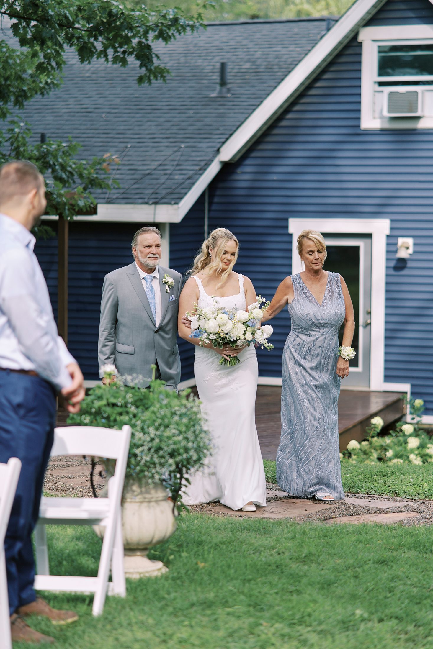 Wedding party seated on white chairs on a lawn in front of a blue house during an outdoor celebration.