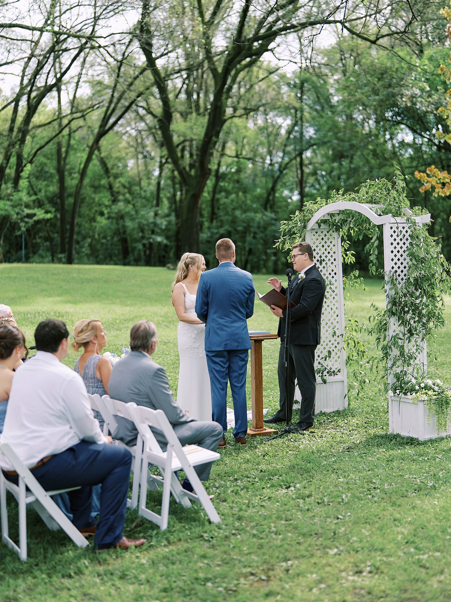 Outdoor wedding ceremony setup with white chairs and archway arranged on a grassy lawn surrounded by trees.