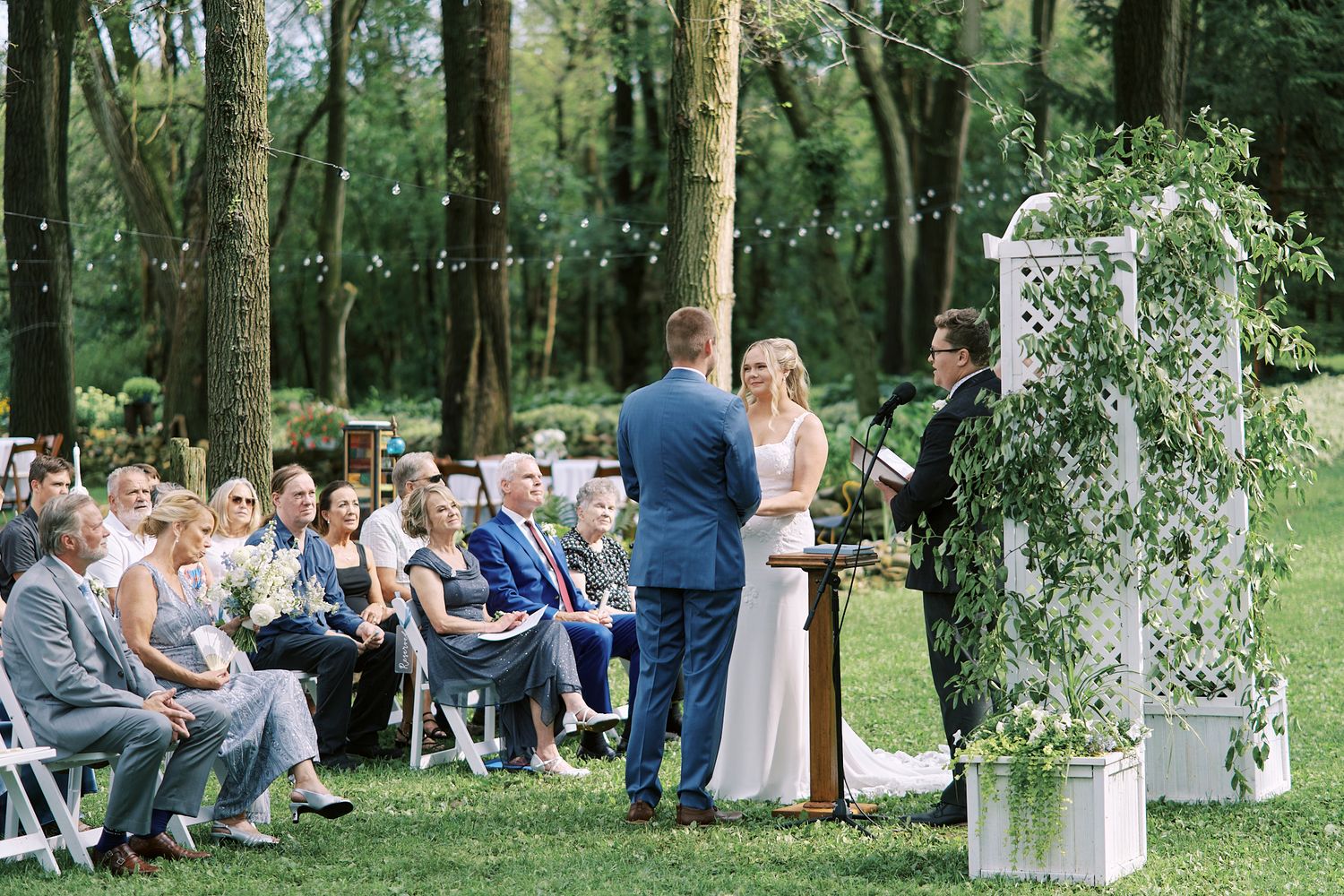 Outdoor wedding ceremony setup in a wooded area with white chairs and decorative arch.