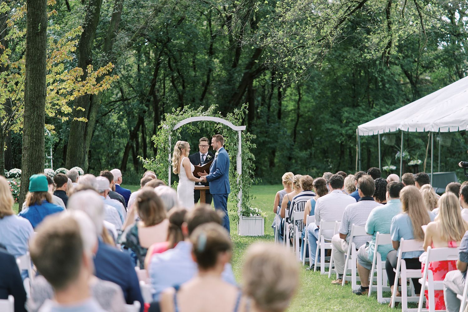 Guests seated in white chairs at an outdoor forest wedding ceremony with natural greenery backdrop.