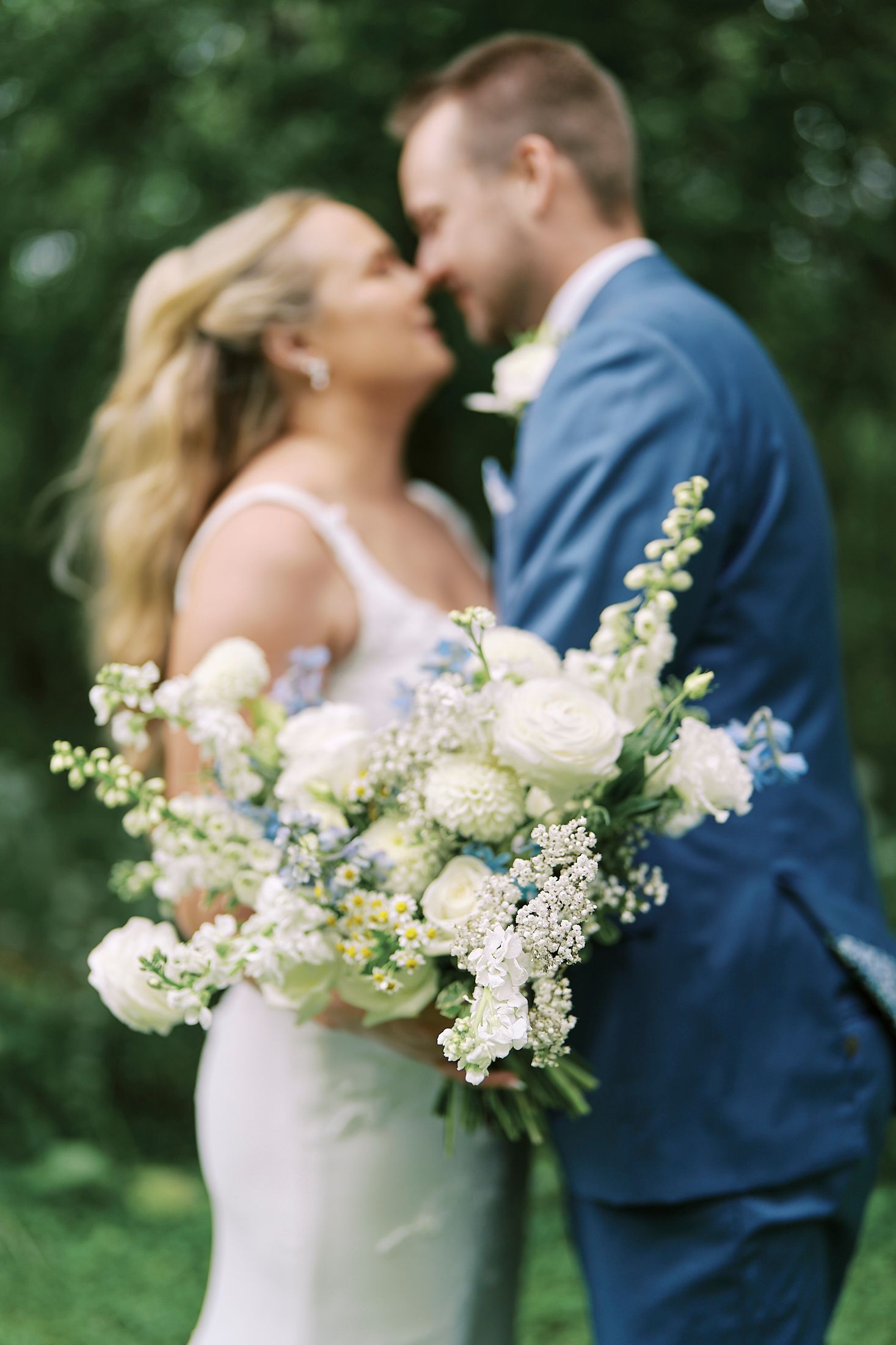 Wedding couple embraces in romantic outdoor setting with white floral bouquet details.