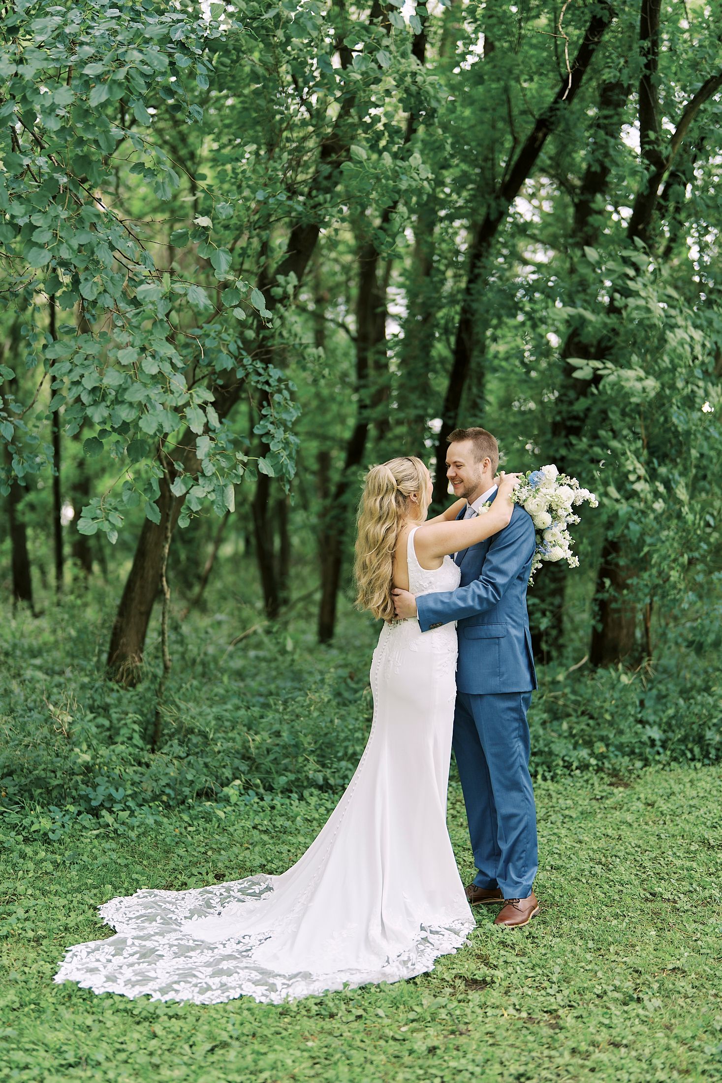 A couple shares a romantic first look moment in a lush green forest setting with a flowing white wedding dress train.