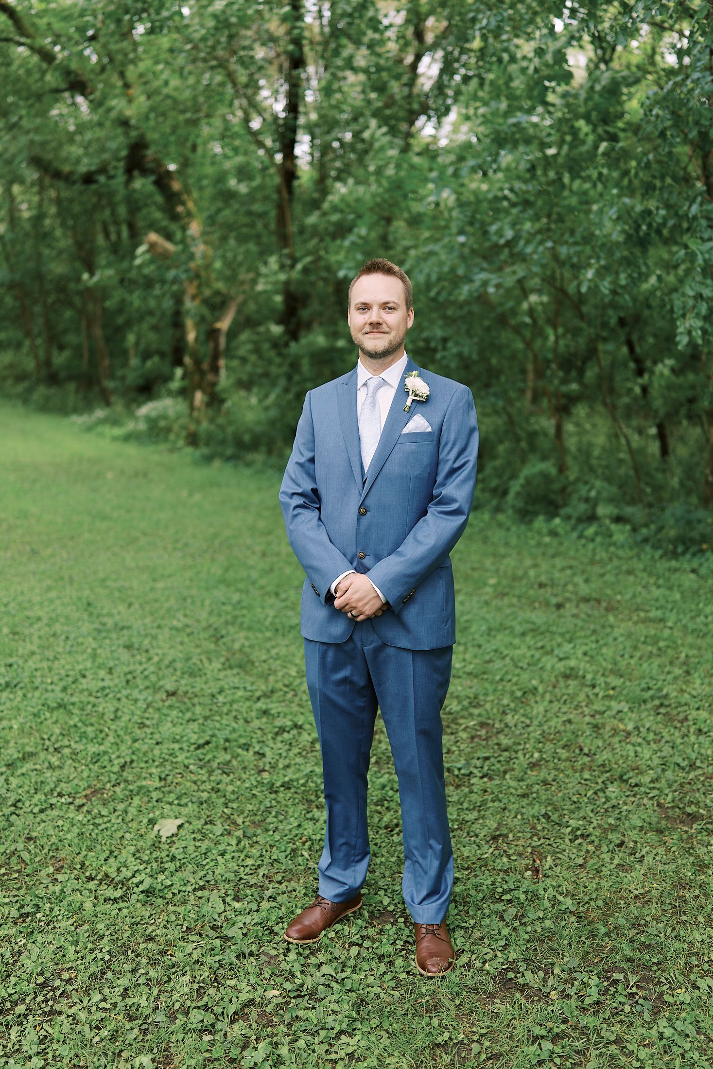 Groom in blue suit and brown shoes poses outdoors on green grass surrounded by trees at outdoor wedding venue.