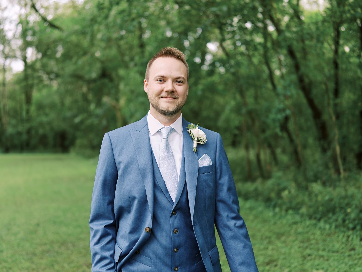 Groom in tailored blue suit stands in green garden setting.