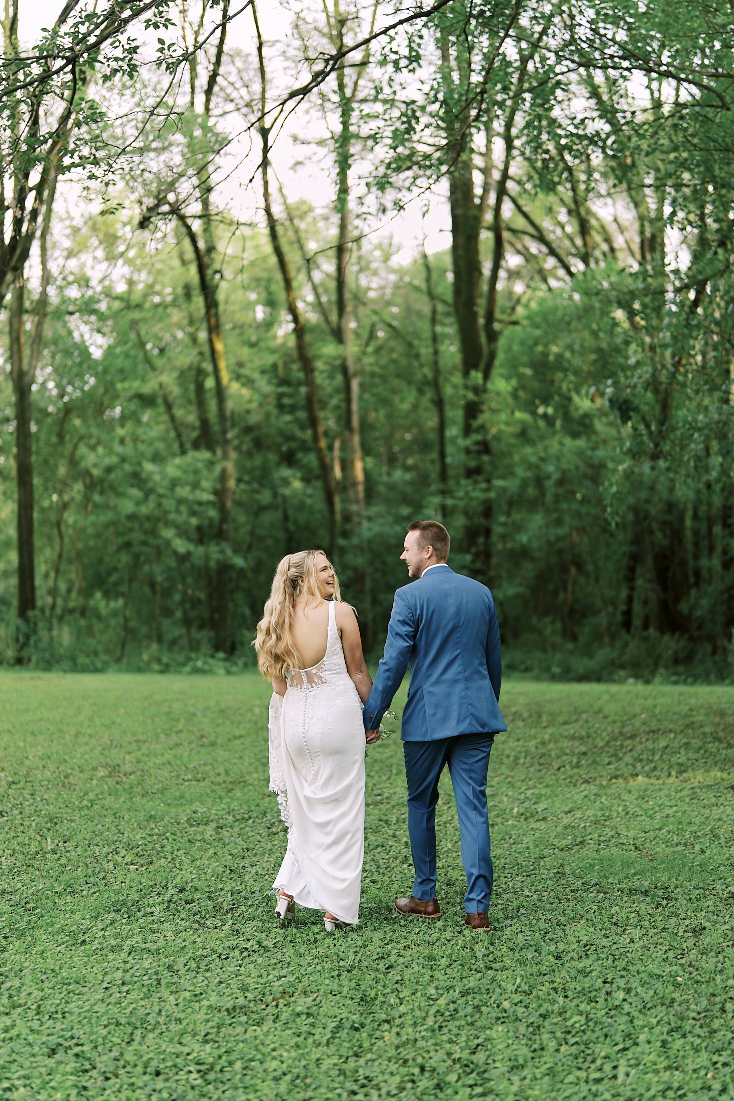 A couple walks hand in hand through a lush green meadow surrounded by trees in a romantic outdoor setting.