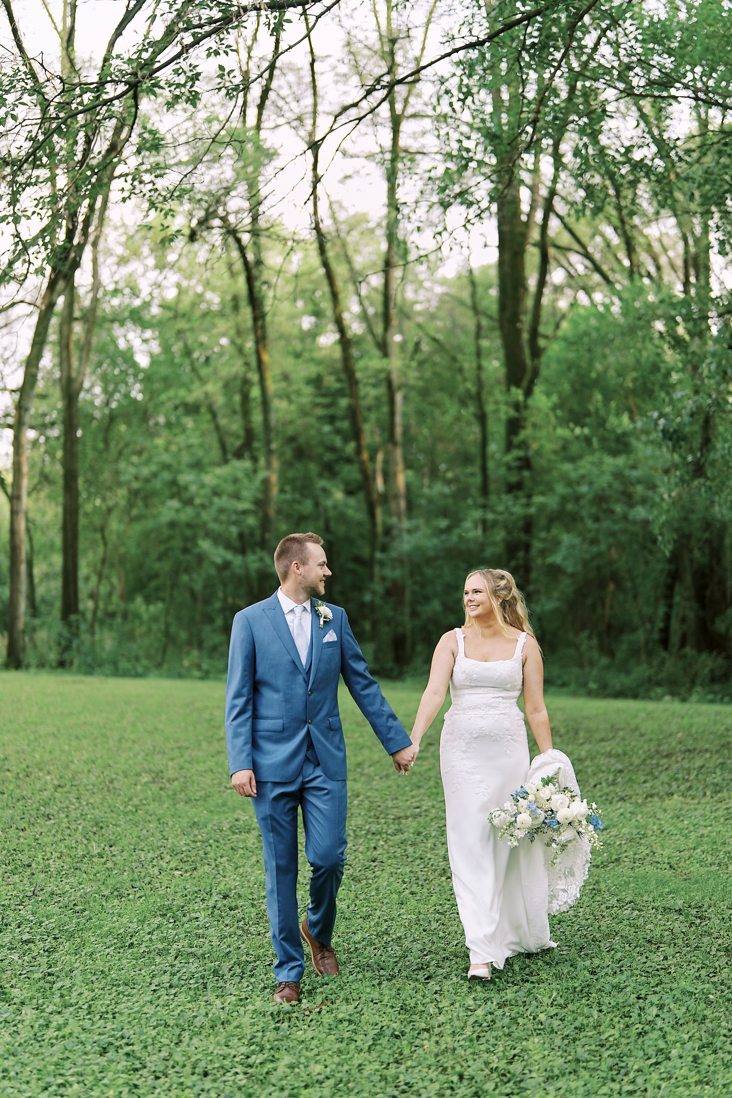 A wedding couple walks hand in hand across a green lawn with a wooded backdrop on their special day.