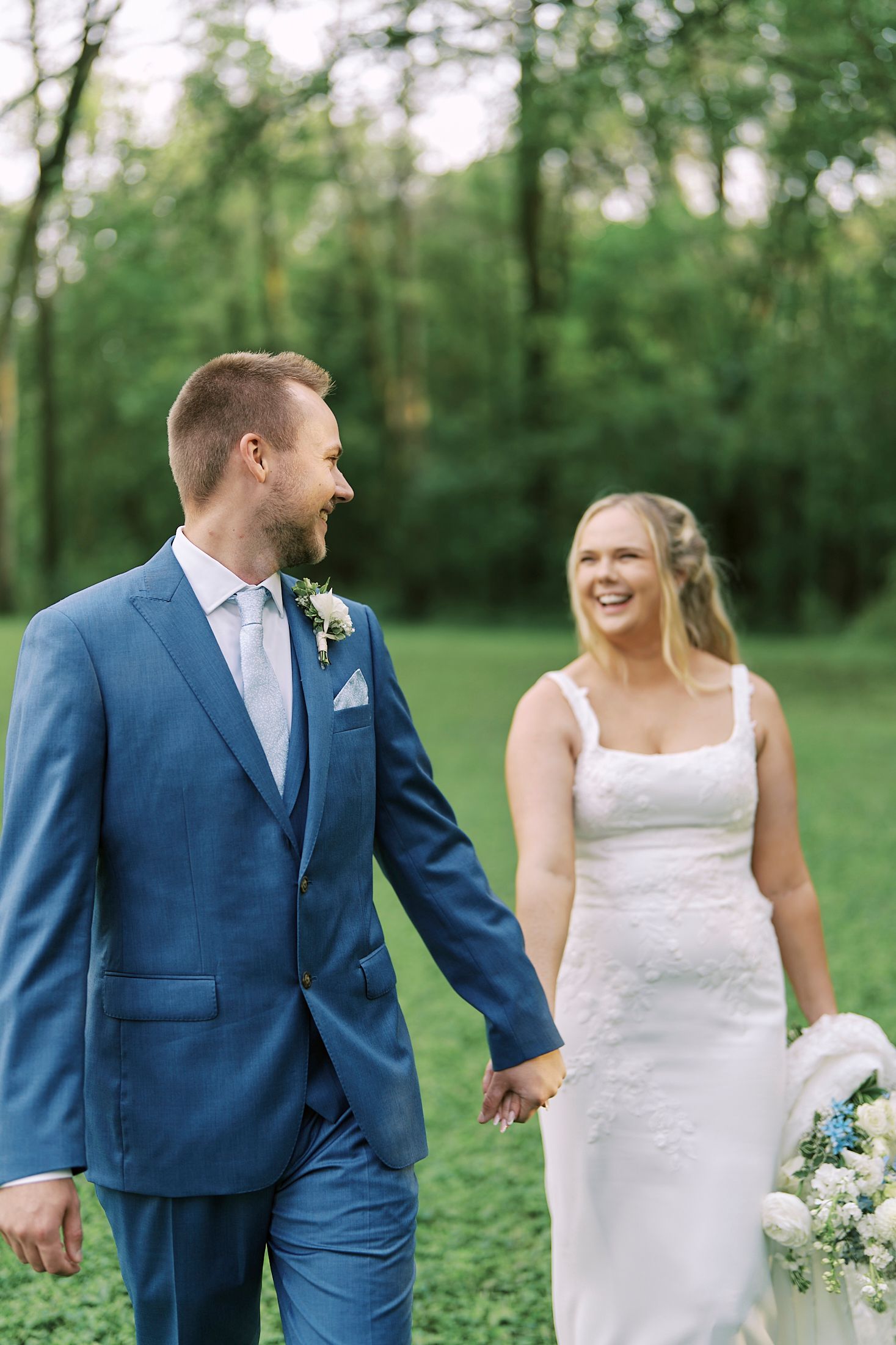 Wedding couple in navy blue suit and white dress walking together in a green garden setting.