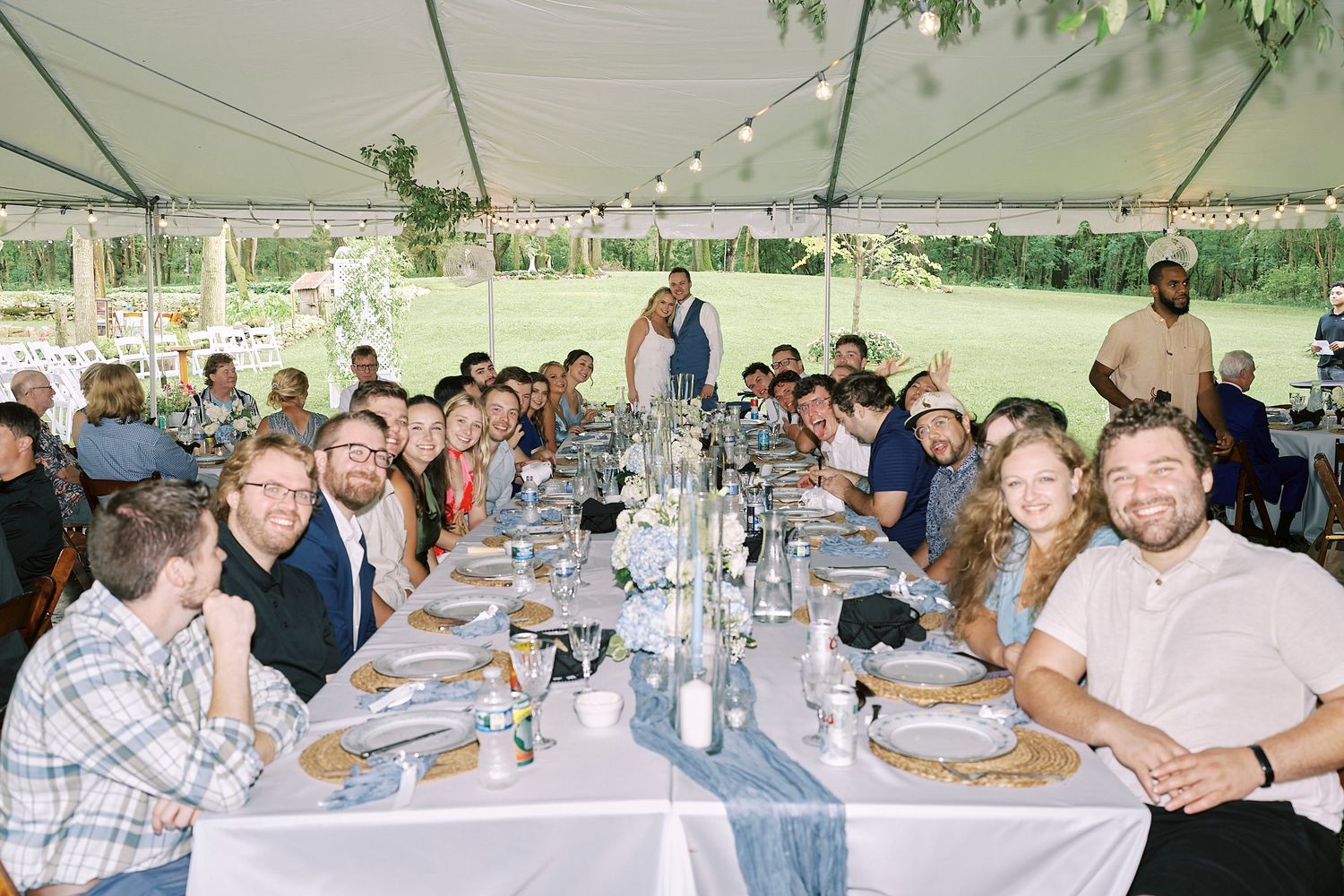 Wedding guests enjoying a lively reception dinner under a white tent with string lights and elegant table settings.