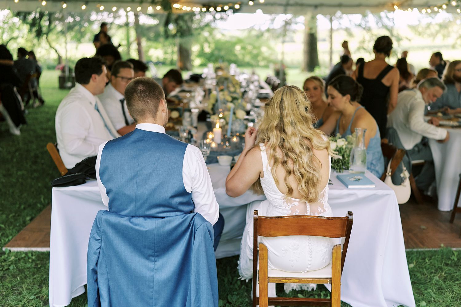 Wedding reception setup at an outdoor venue with multiple tables of guests dining under trees.