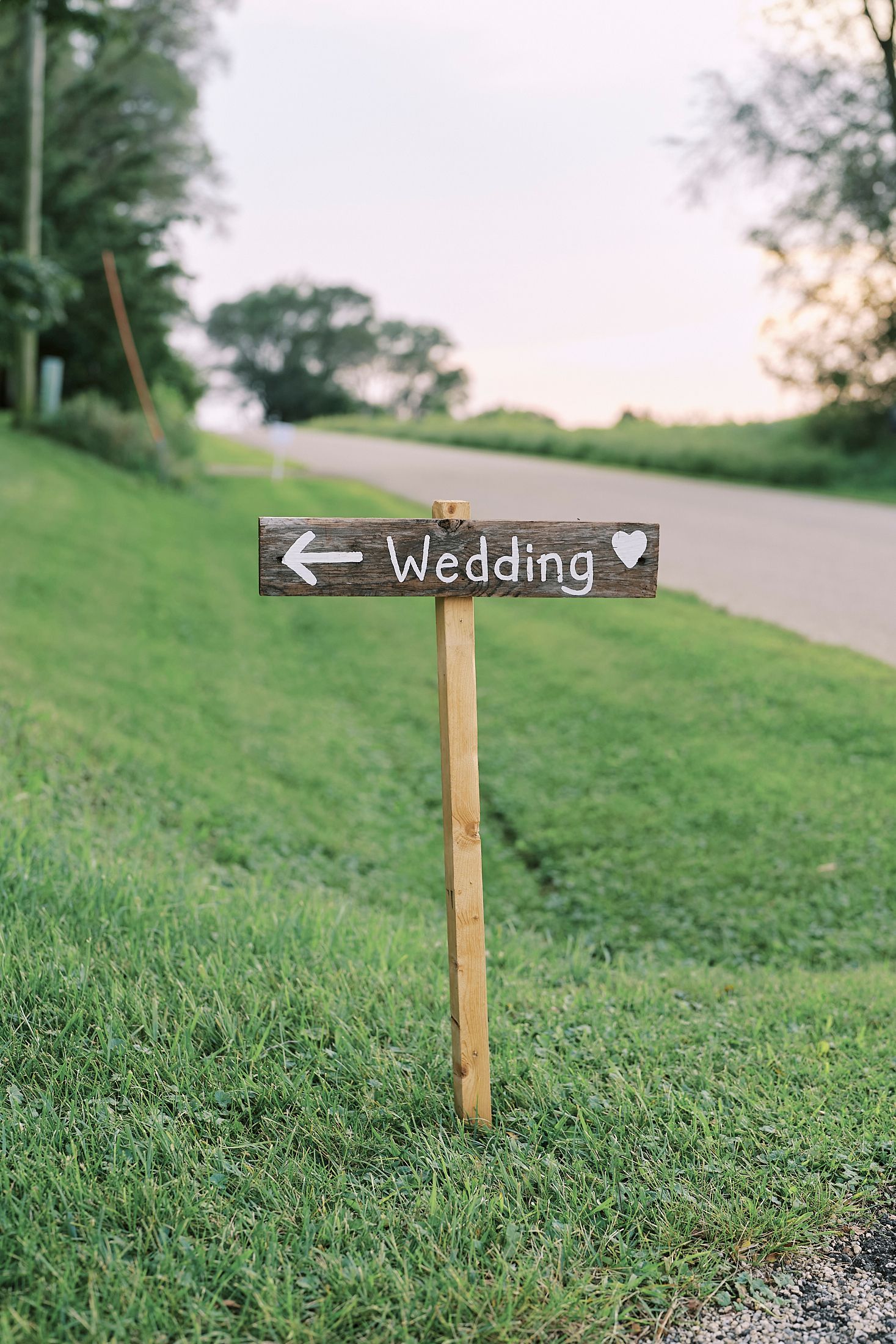 A rustic wooden wedding directional sign with arrow points the way on a grassy hillside path.
