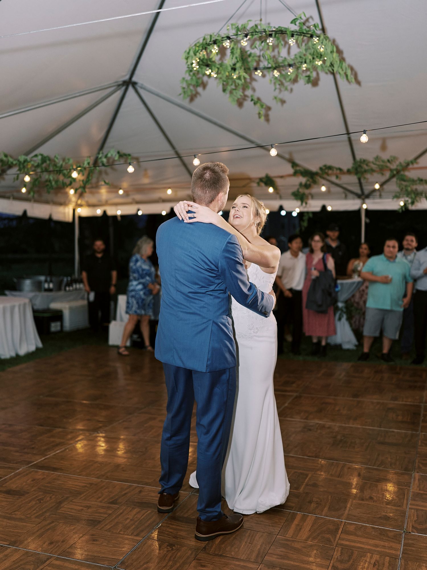 A couple shares a romantic dance moment under a white tent with string lights at an outdoor wedding reception.