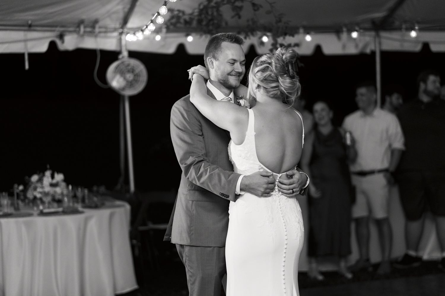 Wedding couple dances intimately on a wooden dance floor under a white reception tent during evening celebrations.