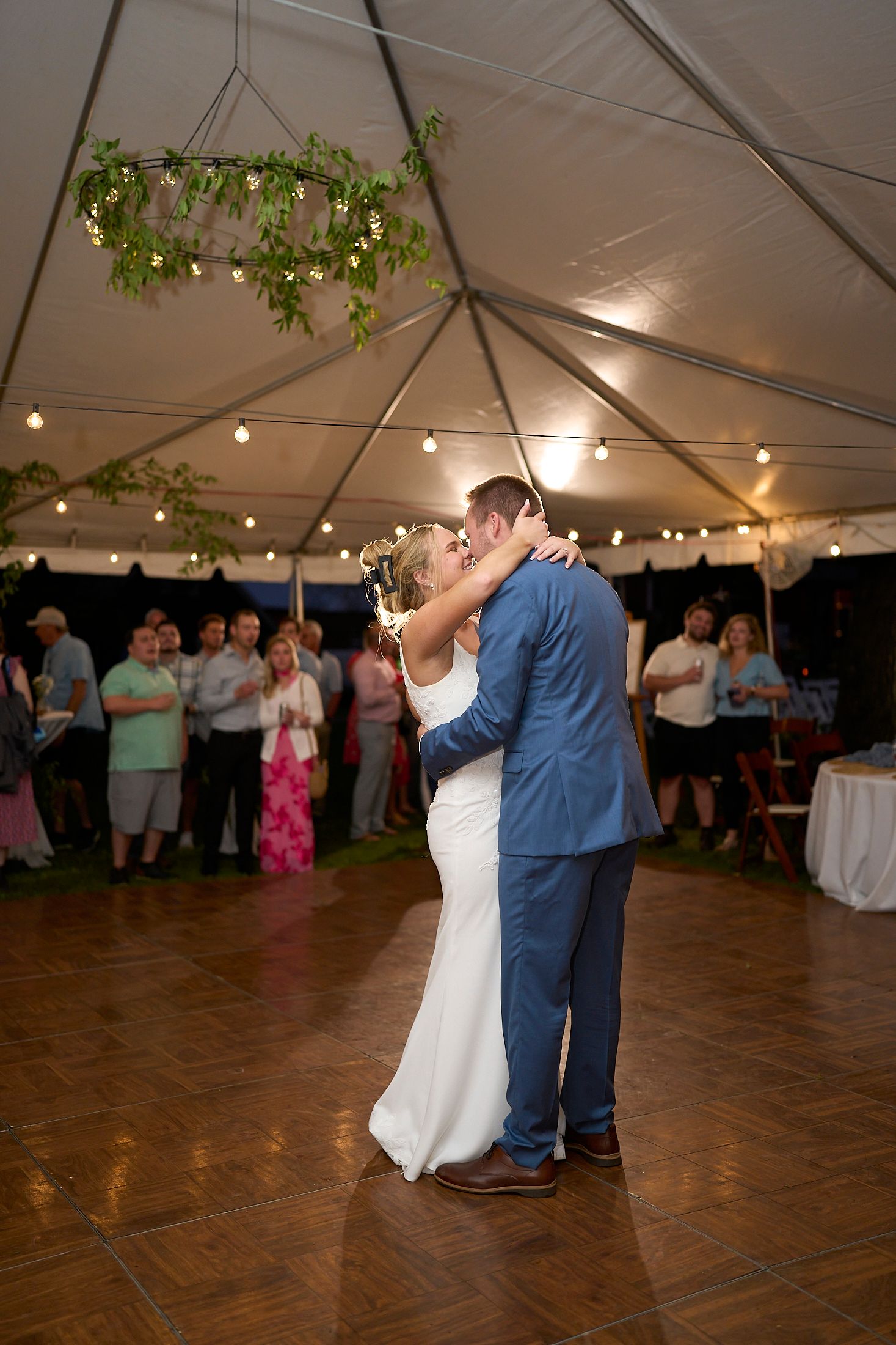 Wedding reception dance floor under tent with string lights and guests celebrating on a wooden floor.