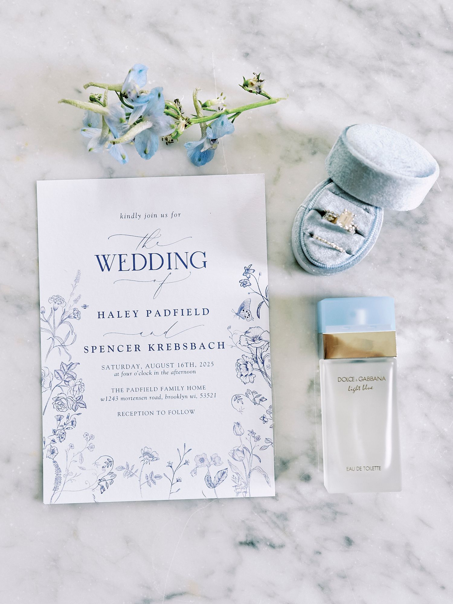 Elegant light blue and white wedding invitation suite with floral designs and matching decorative elements laid out on marble.