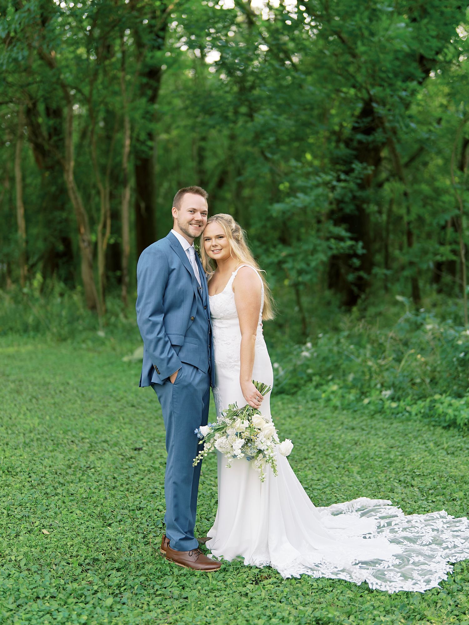 Wedding couple stands together in a lush green forest setting with a flowing white lace dress trailing on the ground.