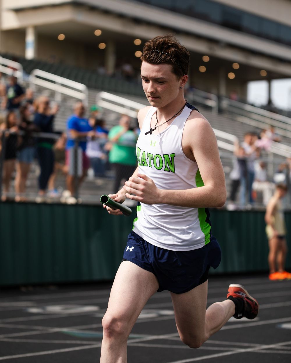 High school track athlete in white and green uniform running on outdoor track during competition while spectators watch.