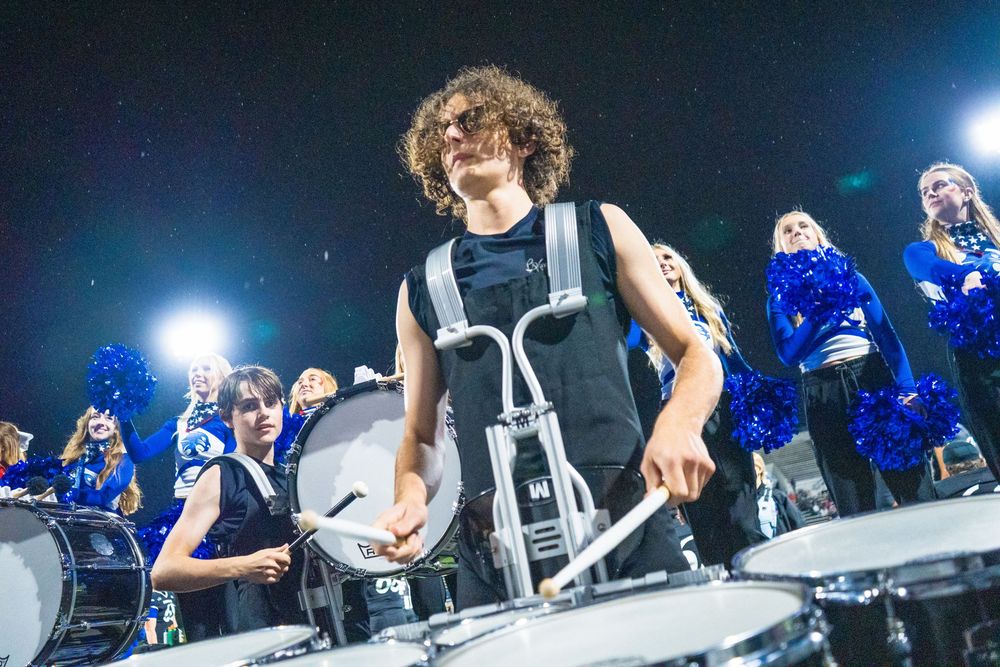 Performer playing drums on stage with colorful blue lighting and audience visible in the background.