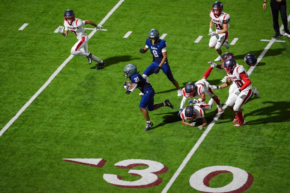 College football players battle for position during an intense play at the 30-yard line on a bright green field.