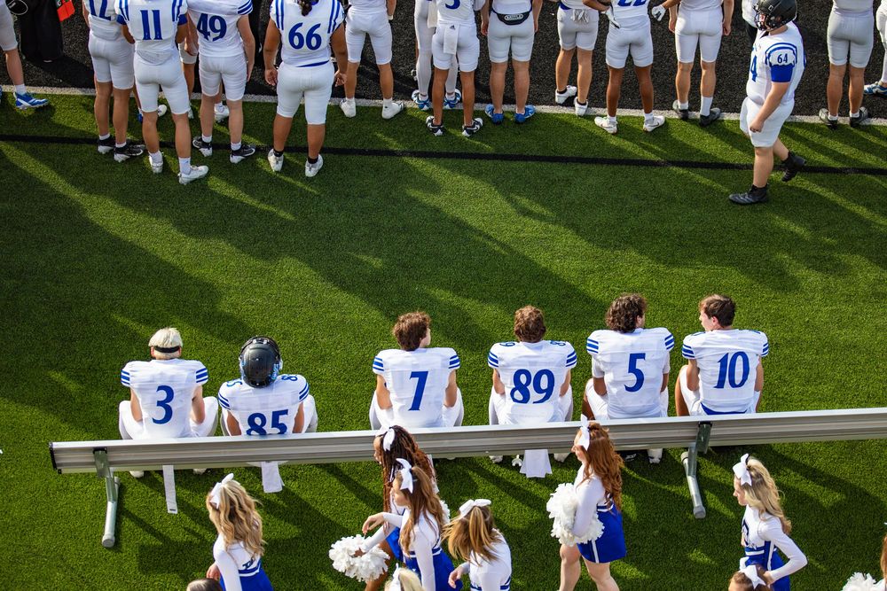 Athletes in white and blue uniforms line up along the sideline of a bright green sports field during a game.